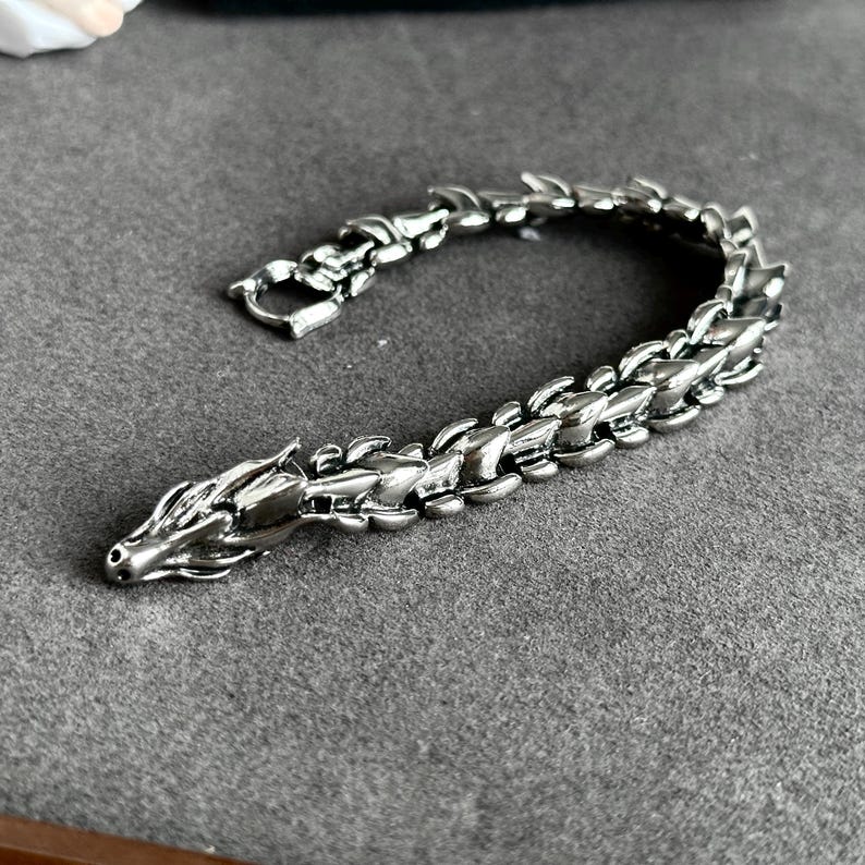 May include: A silver-colored dragon bracelet. The bracelet is made of metal and features a dragon head at one end and a clasp at the other. The dragon's body is segmented, and the bracelet is lying on a gray surface.