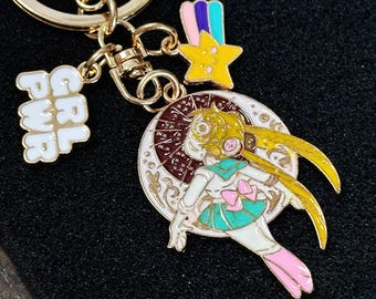 Puede incluir: Un llavero de metal dorado con un dije circular que muestra un personaje de anime en blanco, verde azulado y amarillo, un dije de estrella amarilla con una estela colorida y un dije blanco con el texto GRL PWR en letras doradas.