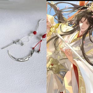 May include: Two silver-toned metal hair accessories featuring intricate sword and crescent moon designs. The sword charm is attached to a white cord with a red bead, while the crescent moon charm hangs from a red cord with a small silver bell.