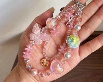 Puede incluir: Un llavero de cuentas con múltiples hilos que presenta un dije de mariposa rosa translúcida, un corazón nacarado, un pequeño dije de caballo mecedora dorado, una cuenta de rosa multicolor y varias cuentas esféricas transparentes, rosas y blancas en una argolla plateada.