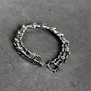 May include: A silver-toned bracelet with a dragon design. The bracelet features a dragon head clasp and a chain-like pattern that resembles a dragon's spine. The bracelet is made of a shiny metal and is displayed on a gray surface.