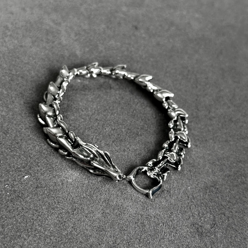 May include: A silver-toned bracelet with a dragon design. The bracelet features a dragon head clasp and a chain-like pattern that resembles a dragon's spine. The bracelet is made of a shiny metal and is displayed on a gray surface.