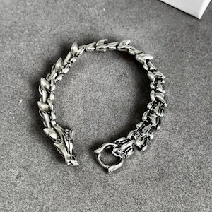 May include: Silver-toned dragon bracelet. The bracelet features a detailed dragon head at one end and a clasp at the other. The links are designed to resemble dragon scales. The bracelet is displayed on a gray surface.