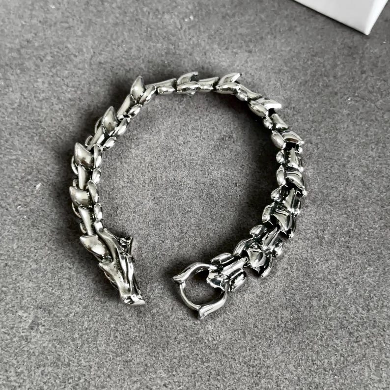 May include: Silver-toned dragon bracelet. The bracelet features a detailed dragon head at one end and a clasp at the other. The links are designed to resemble dragon scales. The bracelet is displayed on a gray surface.