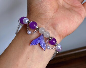 Puede incluir: Una pulsera de cuentas con esferas moradas y blancas, cuentas transparentes y un dije central en forma de cola de sirena morada. El diseño incluye pequeños detalles metálicos en tono plateado y un efecto de cadena drapeada con cuentas.