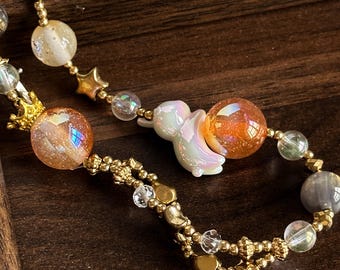 Puede incluir: Una cadena de accesorios con cuentas que presenta dijes dorados de estrellas y coronas, cuentas esféricas iridiscentes en naranja y gris, y una cuenta central blanca iridiscente con forma de conejo. El diseño incorpora pequeños espaciadores dorados.