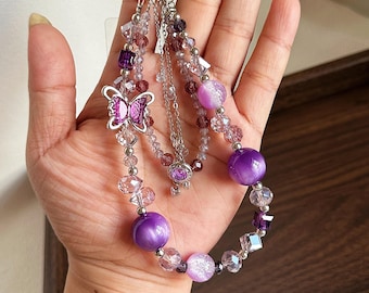 Puede incluir: Un llavero de cuentas con un dije de mariposa de metal plateado, múltiples hilos de cuentas de vidrio facetado moradas y transparentes, y cuentas redondas y lisas de ojo de gato moradas. El accesorio está unido a una argolla de metal plateado.