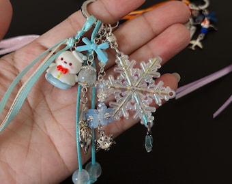 Puede incluir: Un llavero con un dije de muñeco de nieve blanco con lazo rojo, un dije de copo de nieve iridiscente grande, cintas azul claro, cuentas transparentes pequeñas y un lazo azul claro en una argolla de metal plateado con un cristal.