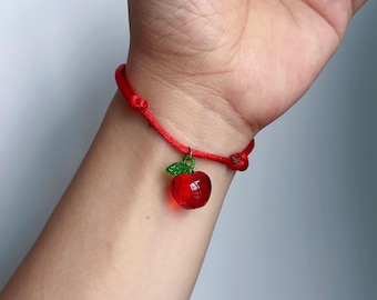 May include: A red cord bracelet featuring a small, translucent red glass apple charm with a tiny green glass leaf attached to the top. The bracelet is secured with a simple knot closure, creating a vibrant and minimalist accessory for the wrist.