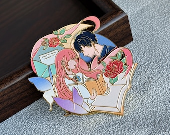 Puede incluir: Un pin de esmalte metálico con la ilustración de una persona de largo cabello rosa leyendo un libro junto a una persona de cabello oscuro. El diseño incluye una mariposa azul, una rosa roja, un sobre verde y contornos de metal dorado.