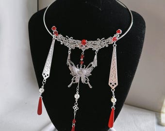 Puede incluir: Un collar tipo gargantilla de metal plateado con una pieza central de filigrana en forma de media luna y un dije de mariposa. El diseño incluye colgantes geométricos plateados, pequeñas perlas blancas y cuentas rojas en forma de gota.