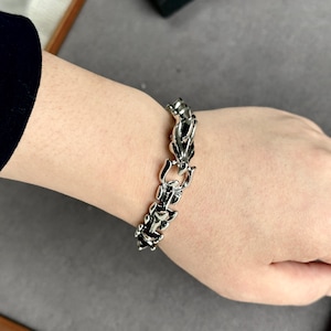 May include: A silver-colored bracelet with a detailed, ornate design. The bracelet is made of interconnected, stylized dragon or animal figures. The clasp is visible, and the bracelet is worn on a wrist, showcasing its intricate craftsmanship and metallic sheen.