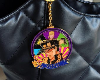 Puede incluir: Un llavero circular de metal dorado con una ilustración en esmalte de colores de personajes de anime, incluyendo una figura central con gorra oscura. El diseño incluye un pequeño ancla y una banda azul con el texto JOJO en dorado.