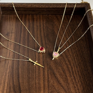 May include: Three gold-tone necklaces with delicate chains. One features a red heart-shaped pendant, another a plain gold sword pendant, and the third a gold sword pendant topped with a pink heart-shaped stone. All pendants have intricate details.