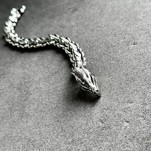 May include: A silver dragon bracelet with a detailed head and a segmented body. The dragon's head is facing right, and the bracelet is on a gray surface. The bracelet is made of metal and has a polished finish.