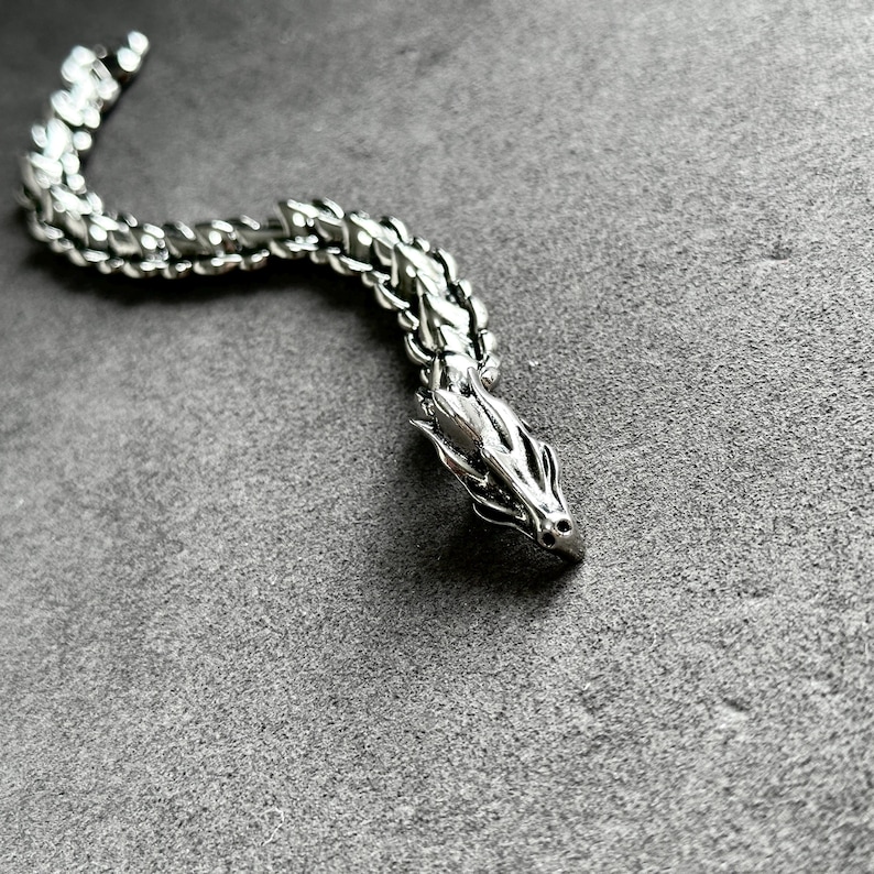 May include: A silver dragon bracelet with a detailed head and a segmented body. The dragon's head is facing right, and the bracelet is on a gray surface. The bracelet is made of metal and has a polished finish.