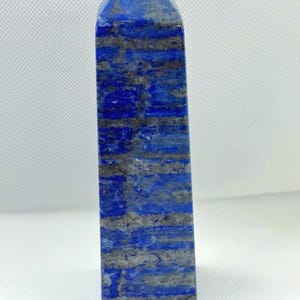 May include: A blue lapis lazuli obelisk with gray veining. The polished stone has a rectangular shape with a pointed top. The stone is standing upright against a white background.