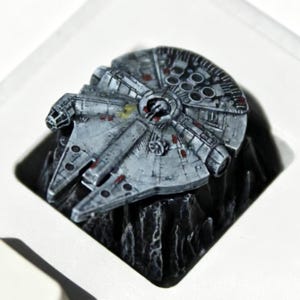 Op de afbeelding: Een gedetailleerde, grijze Millennium Falcon keycap, een miniatuurreplica van het iconische ruimteschip, staat op een zwarte, getextureerde basis. De keycap is geplaatst in een witte vierkant, met een deel van een witte toetsenbordtoets zichtbaar.