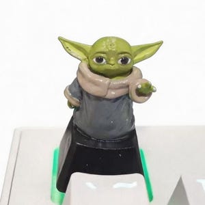 May include: A green and tan figurine of a character from a popular science fiction franchise. The figurine is standing on a white keyboard with green and blue accents. The character is wearing a gray robe and a tan scarf.