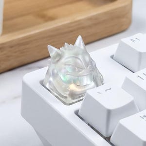 May include: A white mechanical keyboard with a custom keycap shaped like a translucent ghost character. The keycap is placed on the keyboard. A small potted plant and wooden tray are visible in the background. The keyboard keys are white.