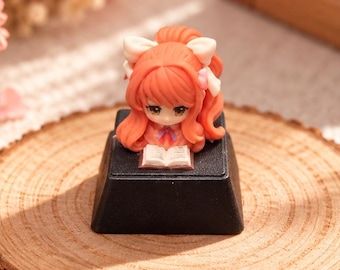 May include: A black mechanical keyboard keycap featuring a small, sculpted figurine of a character with orange hair and white bows, who is holding an open book. The character has large eyes and is positioned on top of the square black keycap base.