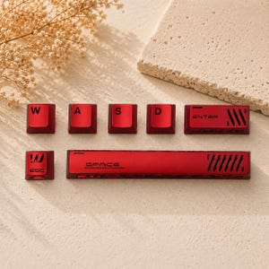 May include: A set of metallic red mechanical keyboard keycaps including W, A, S, D, Enter, Esc, and a long Space bar. Each keycap features black lettering and distinct geometric cutouts or vents, providing a uniform aesthetic for a computer keyboard.