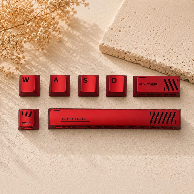 May include: A set of metallic red mechanical keyboard keycaps including W, A, S, D, Enter, Esc, and a long Space bar. Each keycap features black lettering and distinct geometric cutouts or vents, providing a uniform aesthetic for a computer keyboard.