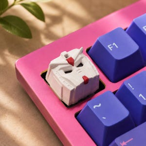 K&ouml;nnte beinhalten: Eine wei&szlig;e, individuell gestaltete mechanische Tastenkappe in Form eines Roboterkopfes mit roten Akzenten, installiert auf einem leuchtend pinkfarbenen Tastaturrahmen neben mehreren violetten Tasten mit wei&szlig; aufgedruckten F1-, Tilde- und Eins-Symbolen.