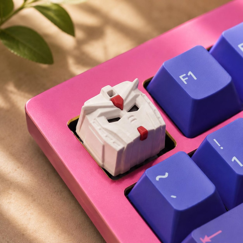 K&ouml;nnte beinhalten: Eine wei&szlig;e, individuell gestaltete mechanische Tastenkappe in Form eines Roboterkopfes mit roten Akzenten, installiert auf einem leuchtend pinkfarbenen Tastaturrahmen neben mehreren violetten Tasten mit wei&szlig; aufgedruckten F1-, Tilde- und Eins-Symbolen.