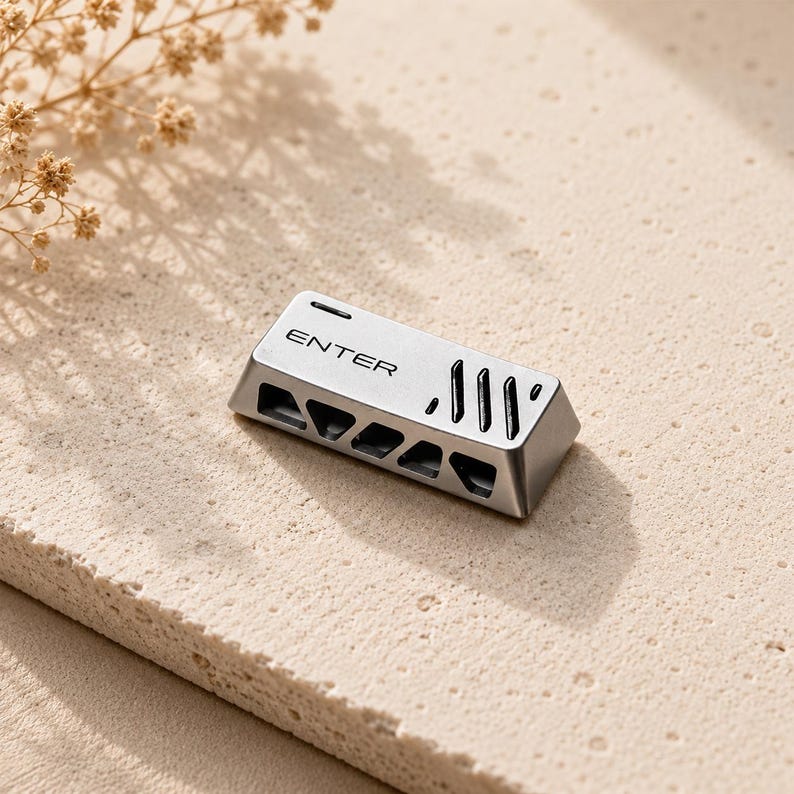 May include: A silver-coloured rectangular metal keycap featuring the word ENTER printed in black. The keycap has a series of triangular cutouts along the front edge and three diagonal black slots on the top surface near the right side.