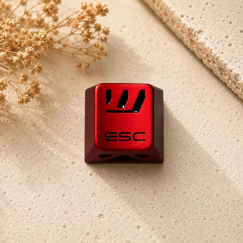 May include: A metallic red mechanical keyboard keycap featuring a cutout design at the top and the text ESC engraved in the centre. The keycap has a smooth, matte finish and a square shape with slightly rounded corners, designed for a keyboard.