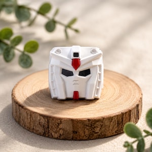 K&ouml;nnte beinhalten: Eine wei&szlig;e Roboter-Kopffigur mit geometrischem Design, bestehend aus kantigen Paneelen, einem zentralen roten Kamm auf der Stirn, einem kleinen roten Kinn-Detail und dunklen, vertieften Augenh&ouml;hlen. Das Objekt hat eine symmetrische Struktur mit zwei Akzenten.