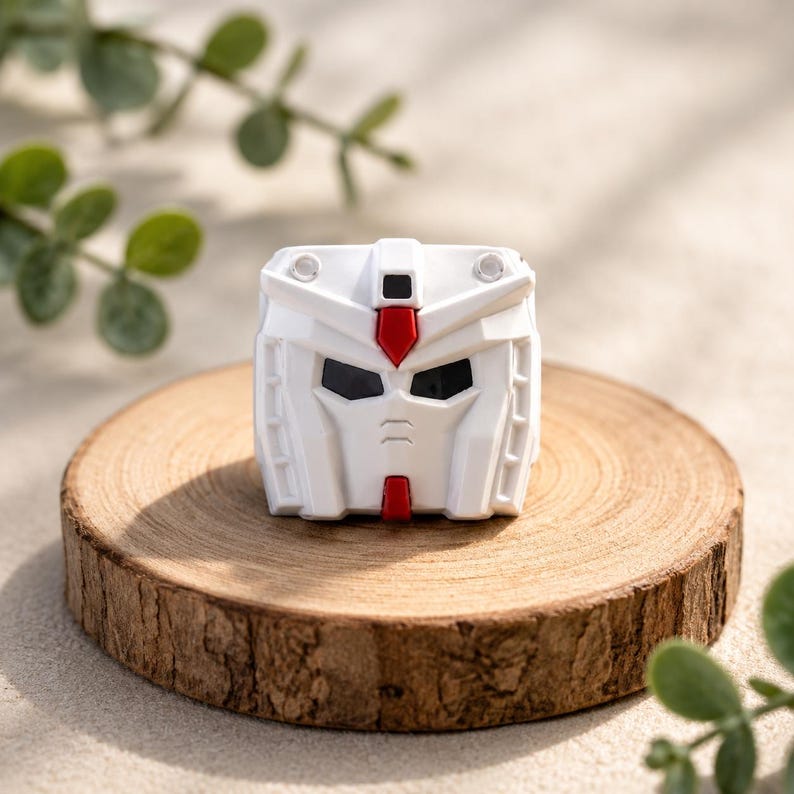 K&ouml;nnte beinhalten: Eine wei&szlig;e Roboter-Kopffigur mit geometrischem Design, bestehend aus kantigen Paneelen, einem zentralen roten Kamm auf der Stirn, einem kleinen roten Kinn-Detail und dunklen, vertieften Augenh&ouml;hlen. Das Objekt hat eine symmetrische Struktur mit zwei Akzenten.