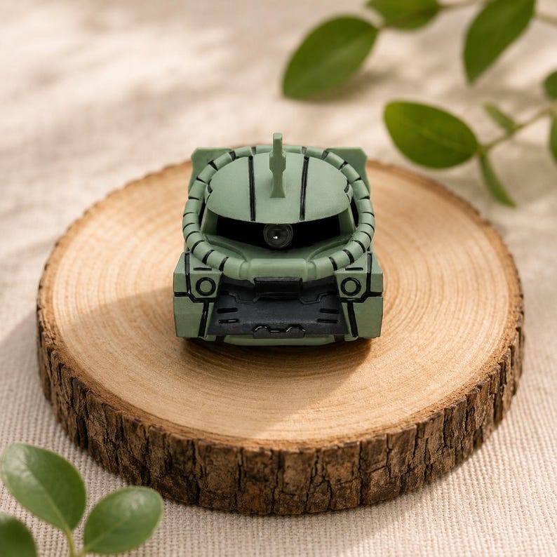 K&ouml;nnte beinhalten: Eine kleine, salbeigr&uuml;ne Kunststofffigur eines Zaku-Mobile-Suit-Kopfes mit zentralem Mono-Eye-Sensor, einer Antenne an der Oberseite und schwarzen Panel-Line-Details. Die Figur hat ein abgerundetes Helmdesign mit einem dunklen, grauen Sockel.