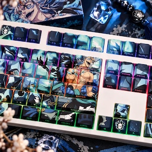 May include: A mechanical keyboard featuring custom keycaps with a blue and black anime-inspired graphic design. The keys display various character illustrations, symbols, and text, including a prominent central image of a shirtless character.