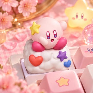 May include: A pink Kirby character figurine sits atop a white cloud-shaped mechanical keyboard keycap. The cloud is decorated with a red heart, a purple star, and a blue heart. A yellow star is attached to Kirby's head, and other pink keycaps are visible.