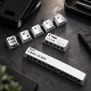 Könnte beinhalten: Ein Set silberner mechanischer Tastaturkappen mit schwarzen Buchstabenaufdrucken und Barcode-Designs. Die Sammlung enthält einzelne Tasten mit der Beschriftung Esc, W, A, S, D, Enter sowie eine lange Leertaste, alle mit metallischem Finish.