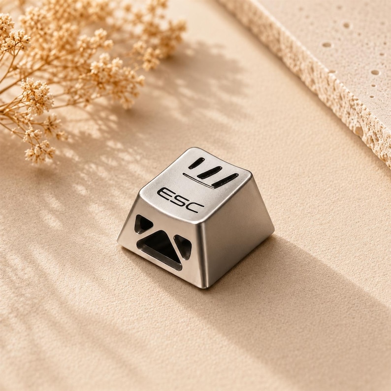 May include: A metallic silver mechanical keyboard keycap featuring the text ESC engraved on the top surface. The keycap has a trapezoidal shape with a cutout design on the front side and additional linear cutouts on the top edge for light.
