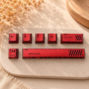 May include: A set of red mechanical keyboard keycaps featuring black accents and symbols. The collection includes an ESC key, a spacebar with the text SPACE, an ENTER key, and four directional arrow keys. Each keycap displays distinct cutouts.