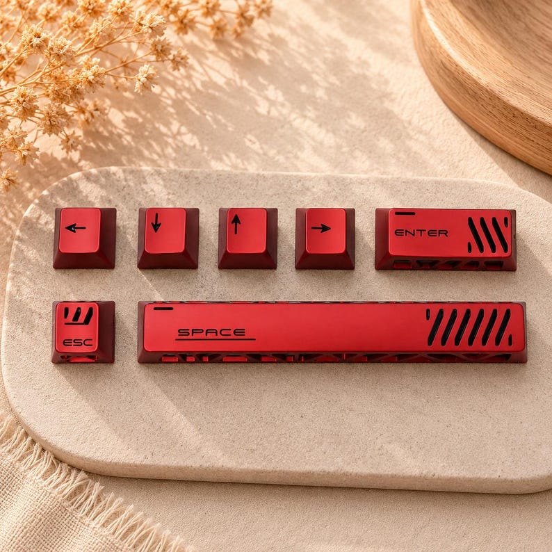 May include: A set of red mechanical keyboard keycaps featuring black accents and symbols. The collection includes an ESC key, a spacebar with the text SPACE, an ENTER key, and four directional arrow keys. Each keycap displays distinct cutouts.