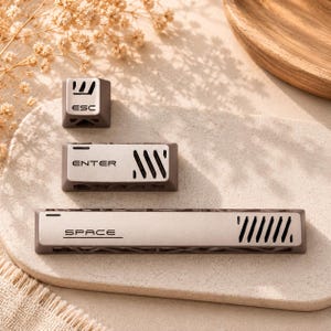 May include: Three mechanical keyboard keycaps in a matte brown and metallic silver finish. The set includes an ESC key, an ENTER key, and a long SPACE bar. Each keycap features black lettering and a series of diagonal ventilation-style cutouts.