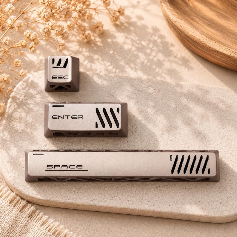 May include: Three mechanical keyboard keycaps in a matte brown and metallic silver finish. The set includes an ESC key, an ENTER key, and a long SPACE bar. Each keycap features black lettering and a series of diagonal ventilation-style cutouts.