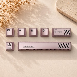 May include: A set of mauve and white mechanical keyboard keycaps including W, A, S, D, ESC, ENTER, and SPACE keys. The keys feature black lettering and decorative diagonal slit patterns on the ENTER and SPACE bars, with a textured base design.