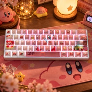 May include: A white mechanical keyboard featuring pink backlit keys with various Kirby character illustrations and the word Kirby printed on the spacebar. The keycaps display custom graphics including small figures, floral patterns, and icons.