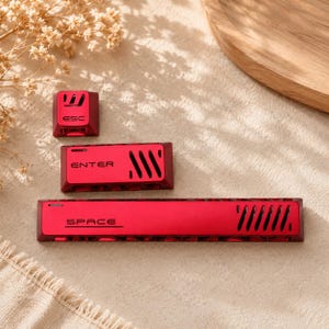 May include: Three red mechanical keyboard keycaps featuring a matte finish and black lettering. The set includes an ESC key, an ENTER key, and a long SPACE bar. Each keycap displays a series of diagonal ventilation slits on the right side.
