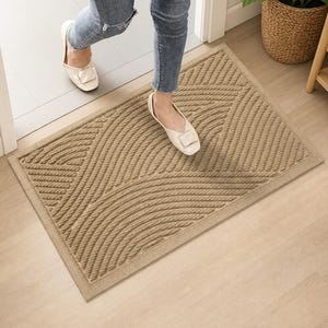May include: Tan rectangular doormat with a textured, geometric design. The mat features curved and angled lines, creating a pattern. The doormat is placed in a doorway, suggesting its use for wiping shoes and preventing dirt from entering a home.