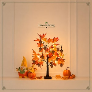 May include: A decorative tabletop tree with orange and yellow autumn leaves, illuminated by small warm lights and adorned with hanging acorn ornaments. Beside it sits a plush gnome figure, a small orange pumpkin, and a larger pumpkin.