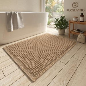 May include: A rectangular, tan waffle-weave bath mat with a fringed edge is placed on a light wood floor. A white bathtub and a folded towel are in the background. A small plant and a wooden shelf with bath products are also visible.