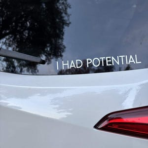 May include: White car with a rear window sticker that reads "I HAD POTENTIAL" in white capital letters. The car's taillight is red and the car is parked outside.
