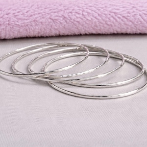 May include: A collection of silver bangles, some with a textured surface, are arranged on a light gray surface. A soft, lavender-colored fabric is in the background. The bangles are simple and elegant.